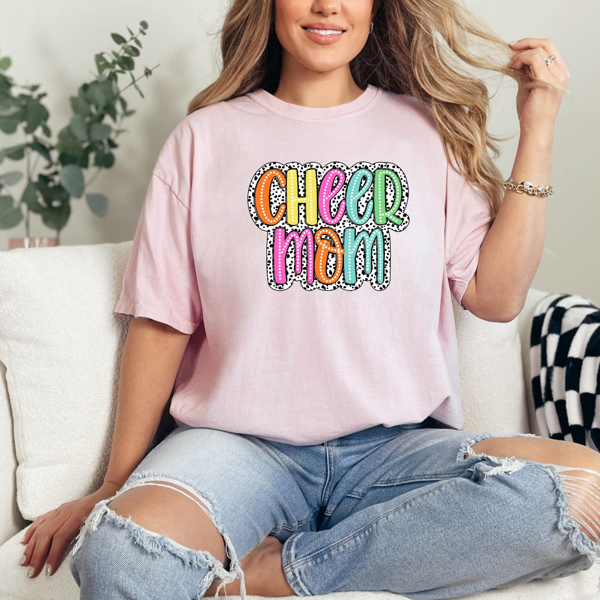 Cheer mom shirt in the color blossom with a bright doodle style font with a background behind the words that appears like black and white dalmatian dots.