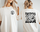 Football mom shirt in ivory with black design.  The front is a small circle corner logo that says footnball mom with a skeleton hand inside.  The back is large and says football mom with a collage of designs including a lighting bolt. football, checkers, hear, and a melting smiley face with a helmet.