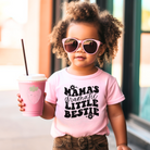 Cute kids graphic tee in pink with a black design.  The text is  a wavy retro design that says "mama's dramatic little bestie" with cute retro flowers on top and below the words.