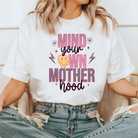 white mom graphic tee with a 90s theme design.  The design says mind your own motherhood in pink, checkered black and white and a yellow melting smiley face.  There are also two small daisies.