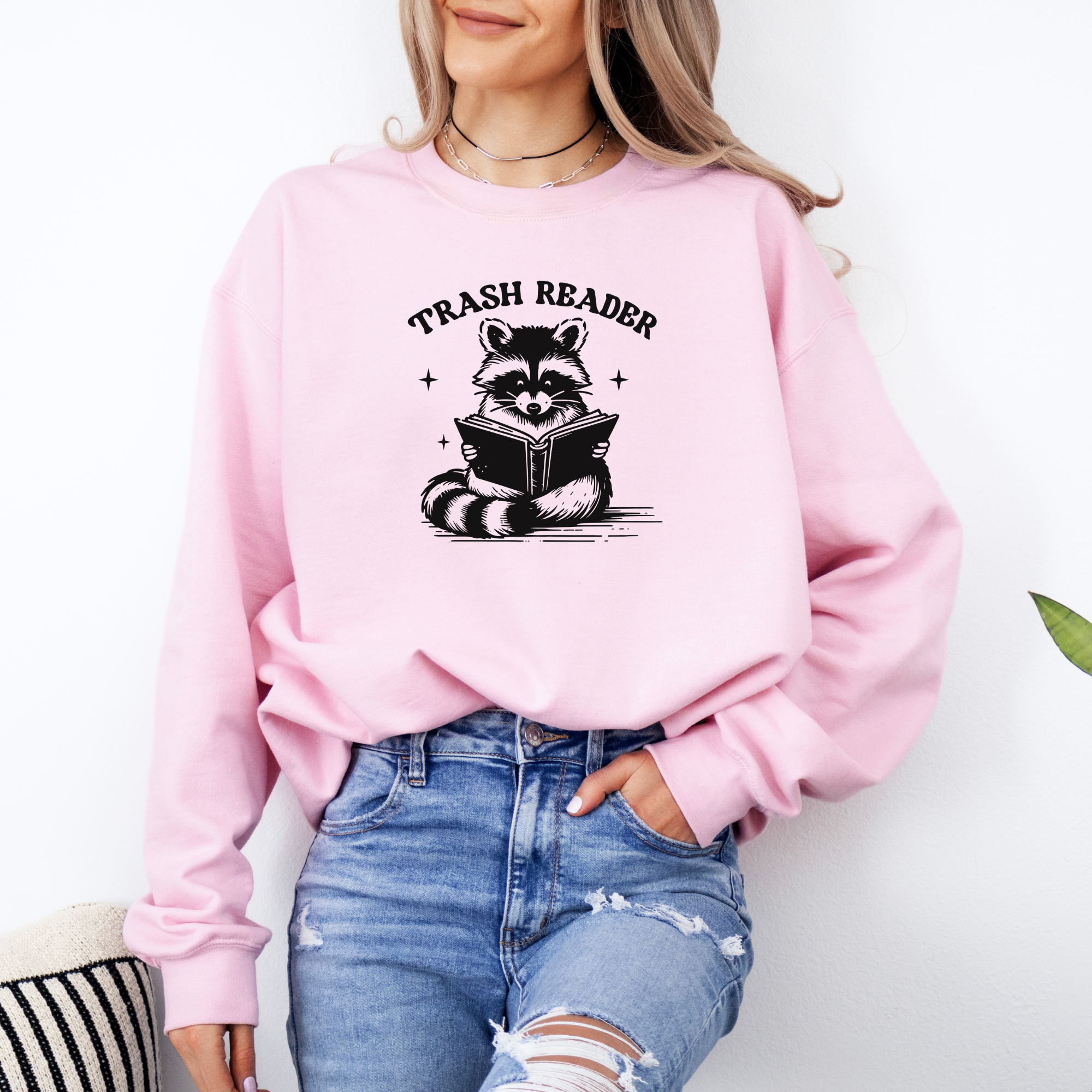 Trash reader with a raccoon reading a book.  The design is black and sweatshirt color is pink.