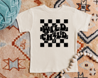 wild child tshirt in the color natural with a black design.  The design is checkered and in the center it says wild child in wavy texy and the I in child is a lightening bolt.