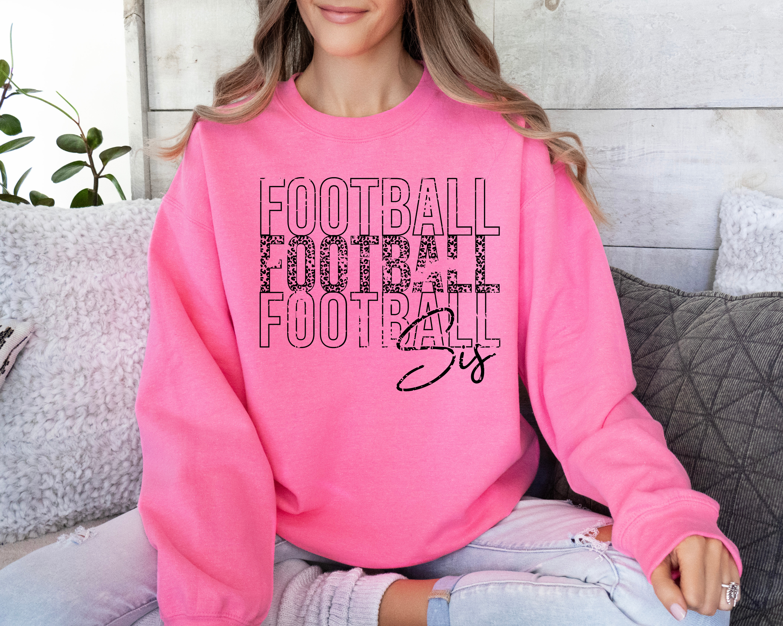 football sister sweatshirt in a safety pink sweatshirt with black design.  The design is faded and stacked saying football in caps in 3 rows.  The middle row is cheetah print lettering.  Below the stack it says sis in cursive.